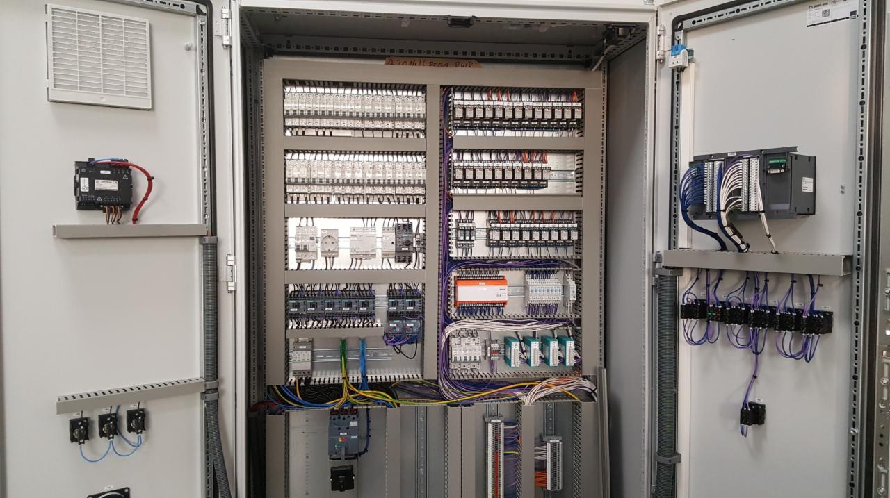 Otwarta szafa sterownicza z licznymi modułami, przewodami i elementami elektrycznymi ułożonymi w uporządkowany sposób. Widoczne różne kolory przewodów i typy złączek.