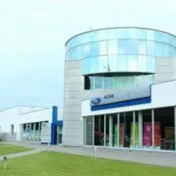 Nowoczesny budynek salonu samochodowego Ford z przeszkloną, cylindryczną wieżą i wyraźnym logo na elewacji. Ujęcie z zewnątrz, z zadbanym trawnikiem i chodnikiem.