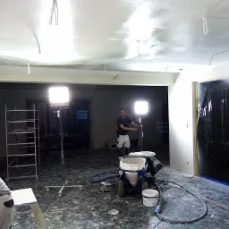 Wnętrze w trakcie prac wykończeniowych: sufit pokryty świeżą warstwą gładzi, widoczna maszyna do nakładania gładzi, osłonięte folią drzwi, oświetlenie budowlane i pracownik w tle.