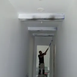 Mężczyzna maluje białą ścianę w korytarzu za pomocą wałka; zabezpieczone folią lampy i klimatyzacja; widoczny pojemnik z farbą i folia ochronna na podłodze.