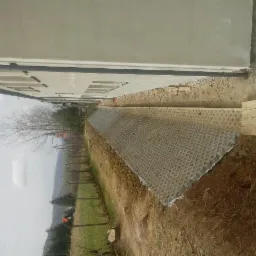 Ułożona ażurowa nawierzchnia wzdłuż ściany budynku, widoczna ziemia i elementy ogrodzenia w tle, pochmurny dzień.