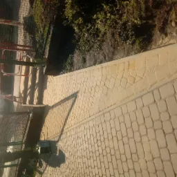 Świeżo ułożona, jasna kostka brukowa w dwóch wzorach: regularny prostokątny i nieregularny mozaikowy, oddzielona szarą obwódką. W tle widoczna siatka ogrodzeniowa i zieleń.