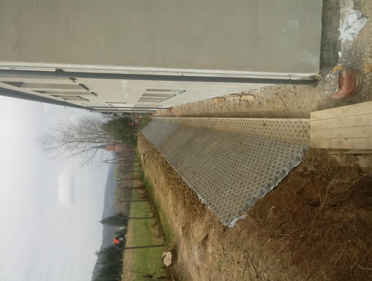 Ułożona ażurowa nawierzchnia wzdłuż ściany budynku, widoczna ziemia i elementy ogrodzenia w tle, pochmurny dzień.