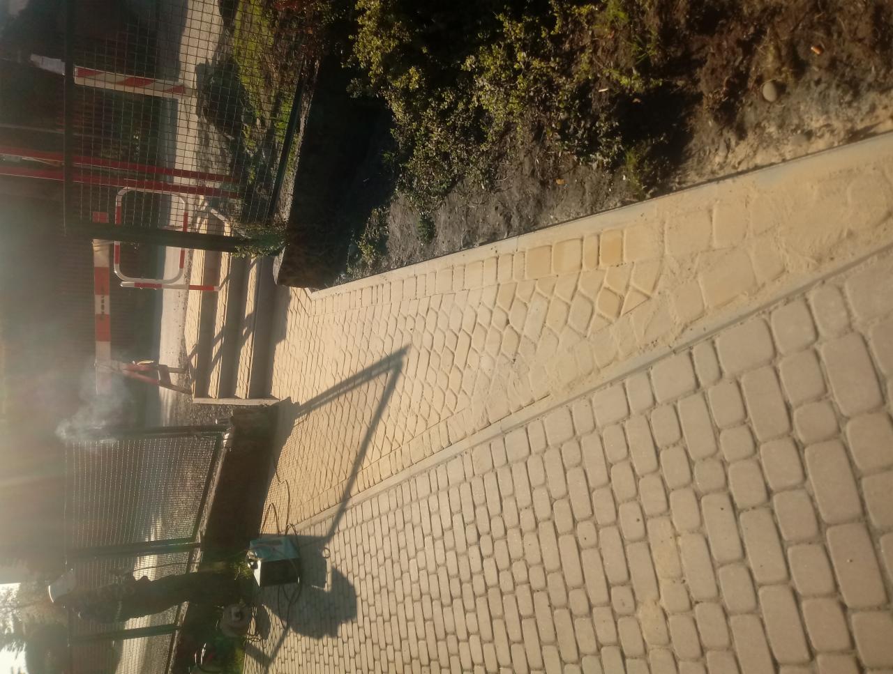 Świeżo ułożona, jasna kostka brukowa w dwóch wzorach: regularny prostokątny i nieregularny mozaikowy, oddzielona szarą obwódką. W tle widoczna siatka ogrodzeniowa i zieleń.