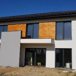 Nowoczesny dom w Stróży z białą elewacją, drewnianymi panelami dekoracyjnymi i dużymi oknami; teren wokół domu przygotowany pod zagospodarowanie.