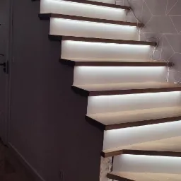 Drewniane schody ze zintegrowanym oświetleniem LED, widoczne przewody, szara ściana z geometrycznym wzorem, drewniana podłoga, boczny widok na schody.