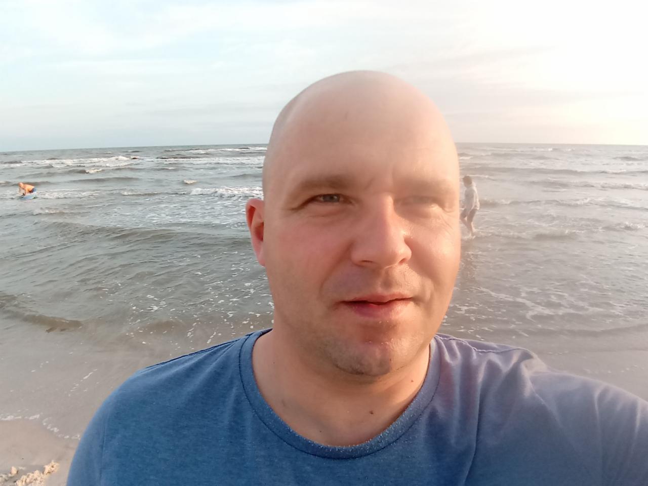 Mężczyzna robi selfie na plaży, w tle morze z falami i osoby w wodzie. Słońce zachodzi, oświetlając twarz mężczyzny. Ubrany w niebieską koszulkę.