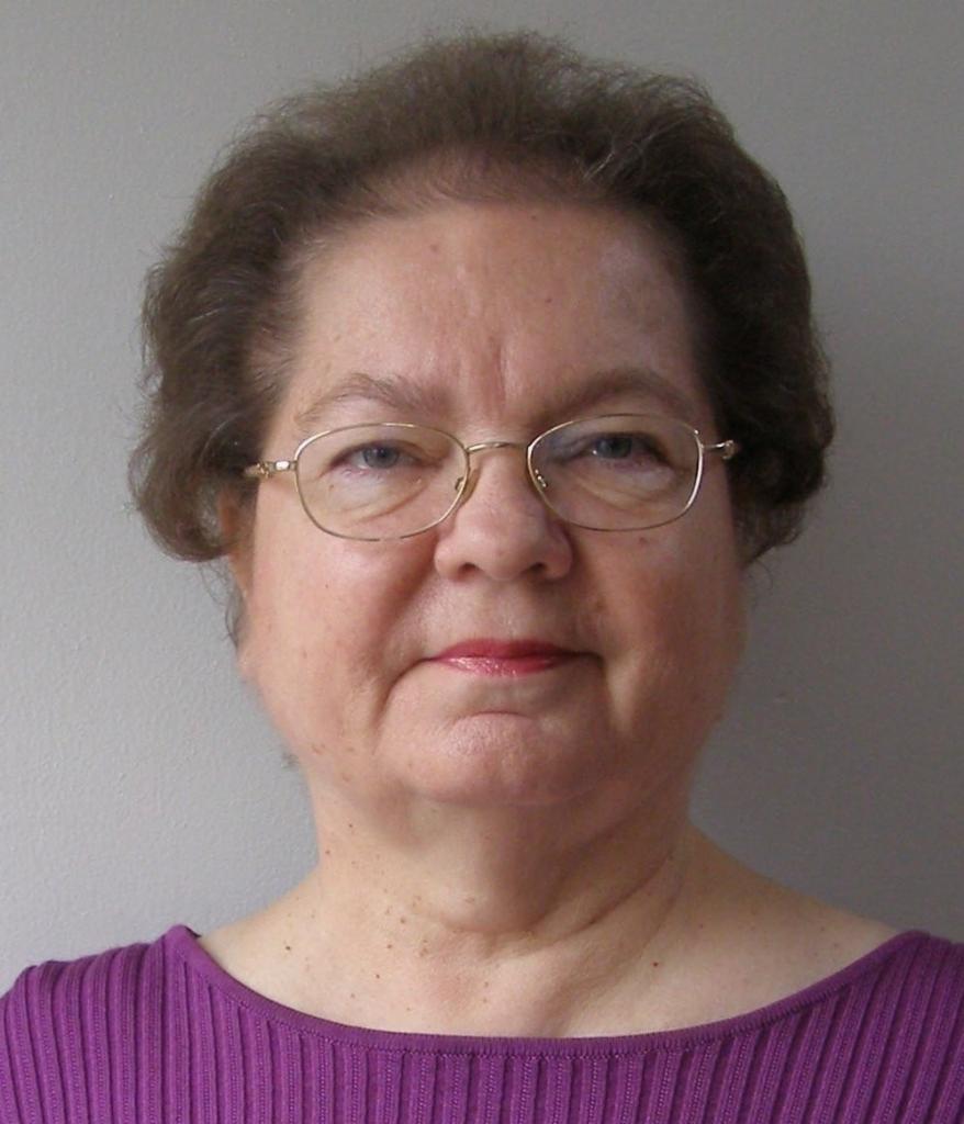 Portret kobiety w okularach i fioletowej bluzce na szarym tle.