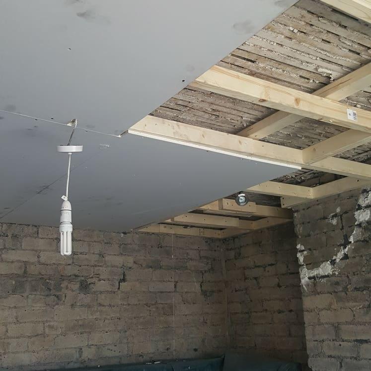 Wnętrze w trakcie prac wykończeniowych: surowa cegła na ścianach, częściowo zamontowany sufit podwieszany z widoczną drewnianą konstrukcją i wiszącą żarówką.