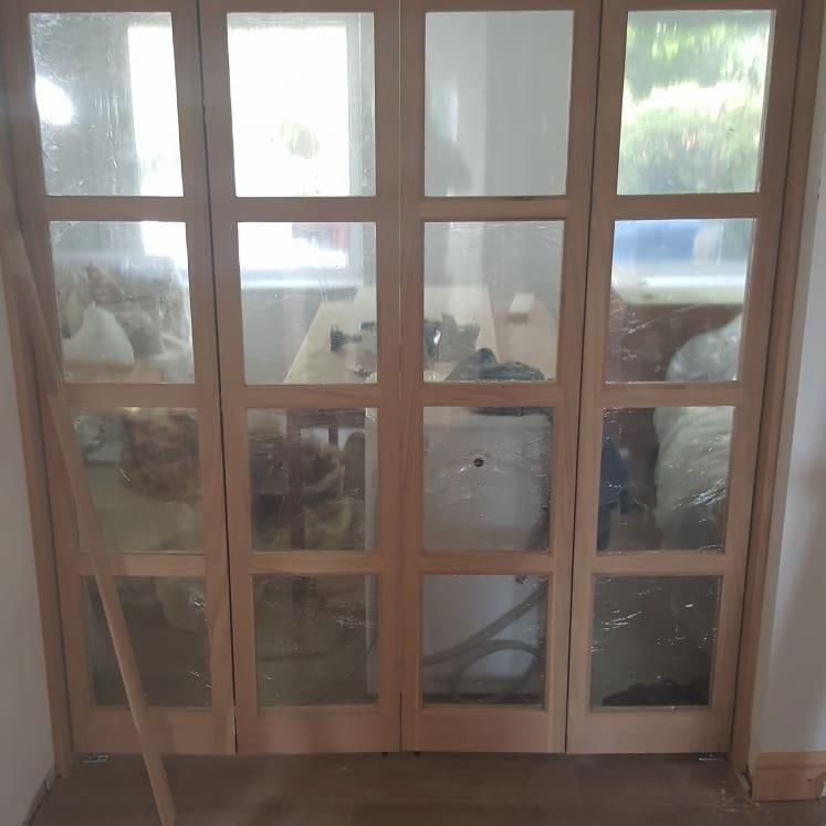 Czteroskrzydłowe, drewniane drzwi z szybami w trakcie montażu, zabezpieczone folią ochronną, widoczne narzędzia i materiały budowlane w tle.