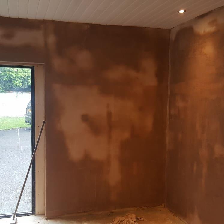 Ściana w trakcie tynkowania, widoczne niedociągnięcia i mokre plamy, sufit z białych paneli, widok na zewnątrz przez okno.