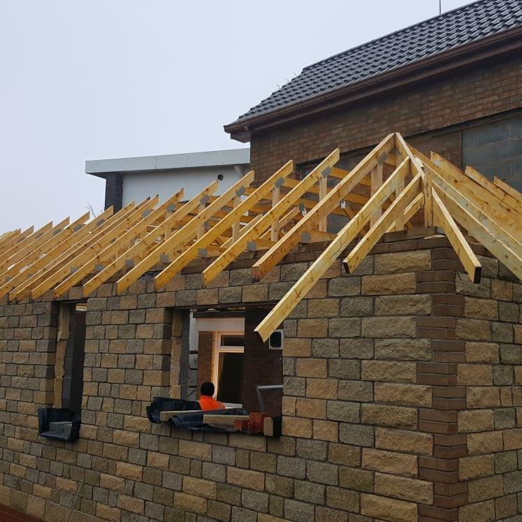 Budowa domu z kamienia: widok na wznoszone ściany z oknami i drewnianą konstrukcją dachu, w tle fragment istniejącego budynku z ciemną dachówką.