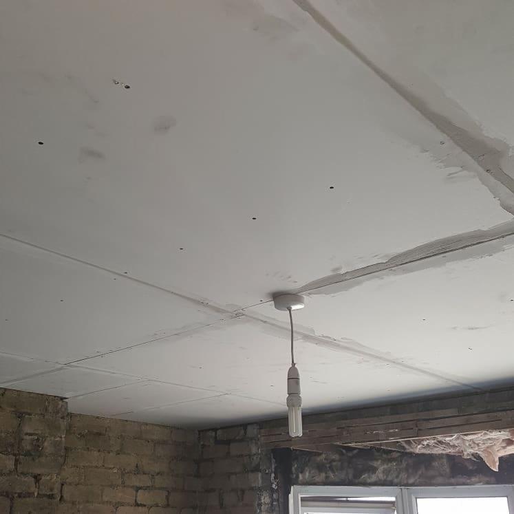 Niedokończony sufit z płyt gipsowo-kartonowych, widoczne szpachlowane łączenia, wisząca żarówka i fragment ściany z cegły.