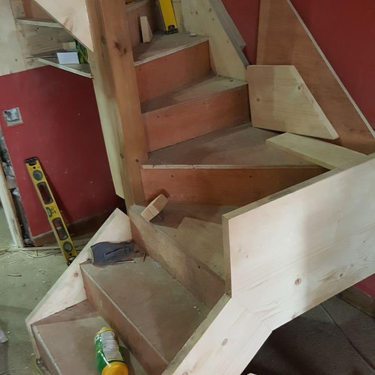 Drewniane schody w trakcie budowy, widoczne elementy konstrukcyjne, poziomica i narzędzia, surowy stan wykończenia, czerwona ściana w tle.