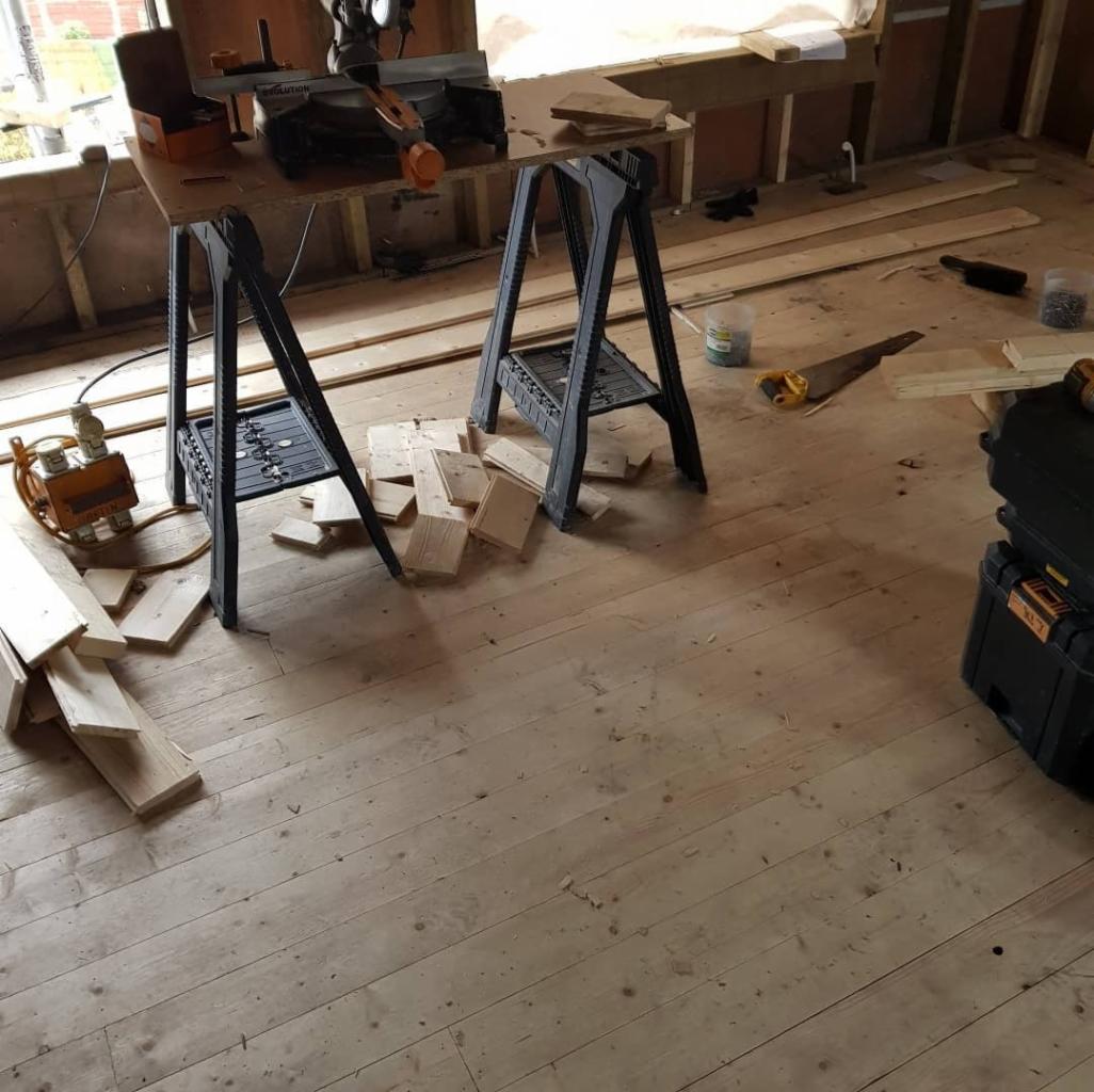 Fragment pokoju w trakcie remontu z widocznymi deskami podłogowymi, stojakami, narzędziami i skrzynką narzędziową, ukazujący etap układania parkietu.