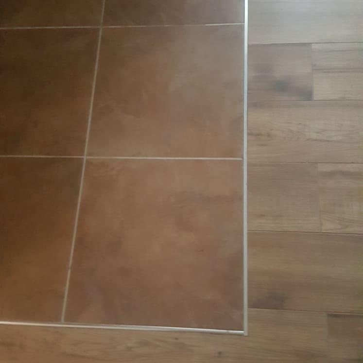 Połączenie brązowych płytek podłogowych z drewnianą podłogą, wykończone srebrną listwą przypodłogową.