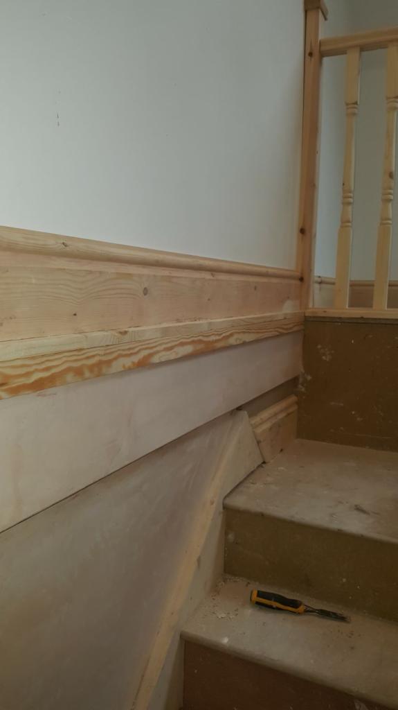 Montaż drewnianej boazerii na ścianie przy schodach, widoczne niedoskonałości w szpachlowaniu i narzędzie pracy - dłuto na stopniu.