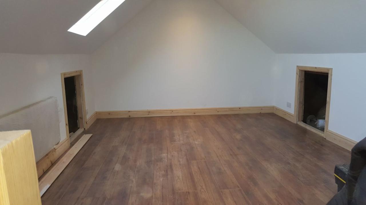 Poddasze z nową, brązową podłogą imitującą drewno, świeżo pomalowane ściany na biało, niskie drzwi i okno dachowe, listwy przypodłogowe z jasnego drewna.