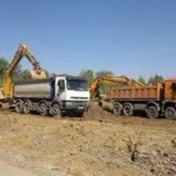 Dwa wywrotki i dwie koparki podczas prac ziemnych na placu budowy, widoczne drzewa w tle, błękitne niebo.