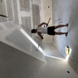 Hetman Bud - Mężczyzna w tank topie maluje ścianę wałkiem w pomieszczeniu z płyt kartonowo-gipsowych, widoczna drabina i kuweta z farbą.