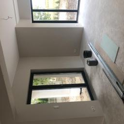 Hetman Bud - Wnętrze w trakcie prac wykończeniowych: ściany przygotowane pod malowanie, widoczne profile, płyta gipsowo-kartonowa, wiadro i kanister na podłodze, duże okna z czarnymi ramami.