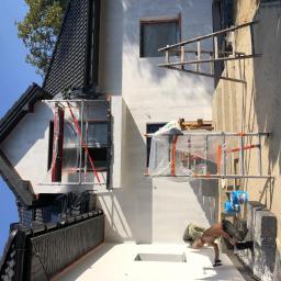 Hetman Bud - Prace wykończeniowe na elewacji domu jednorodzinnego: mężczyzna szpachluje ścianę przy użyciu pacy, obok stoją puszki z farbą, okna zabezpieczone folią, widoczne rusztowania.