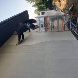 Hetman Bud - Dwaj pracownicy nakładają tynk na elewację budynku jednorodzinnego, widoczna drabina zabezpieczona folią i fragment dachu.