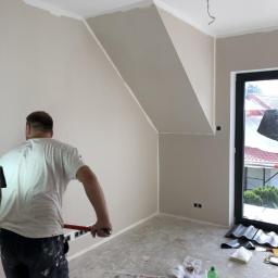 Hetman Bud - Pracownik przygotowuje ścianę w pomieszczeniu ze skosami, widoczne narzędzia i materiały budowlane, ślady po szpachlowaniu i malowaniu, gazeta na podłodze.