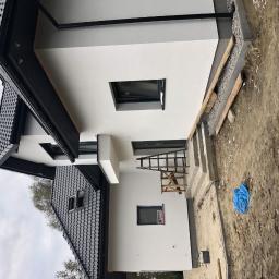 Hetman Bud - Nowoczesny dom z białą elewacją, czarną dachówką i oknami, widoczna drabina oparta o ścianę, teren wokół domu w trakcie prac wykończeniowych.