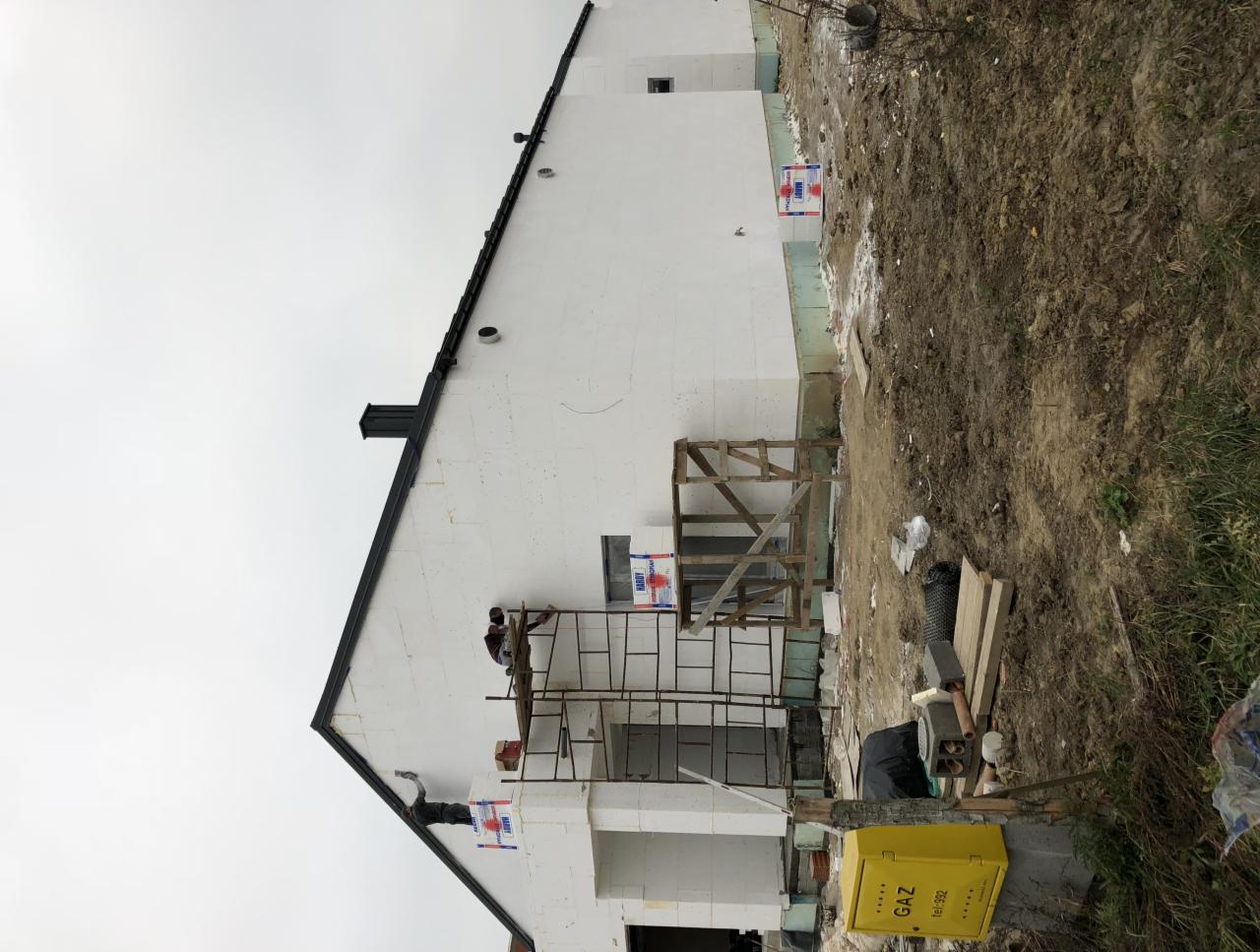 Dwóch pracowników na rusztowaniu podczas nakładania izolacji termicznej na ścianę domu jednorodzinnego. Widoczna żółta skrzynka gazowa, teren budowy i nieotynkowana elewacja.