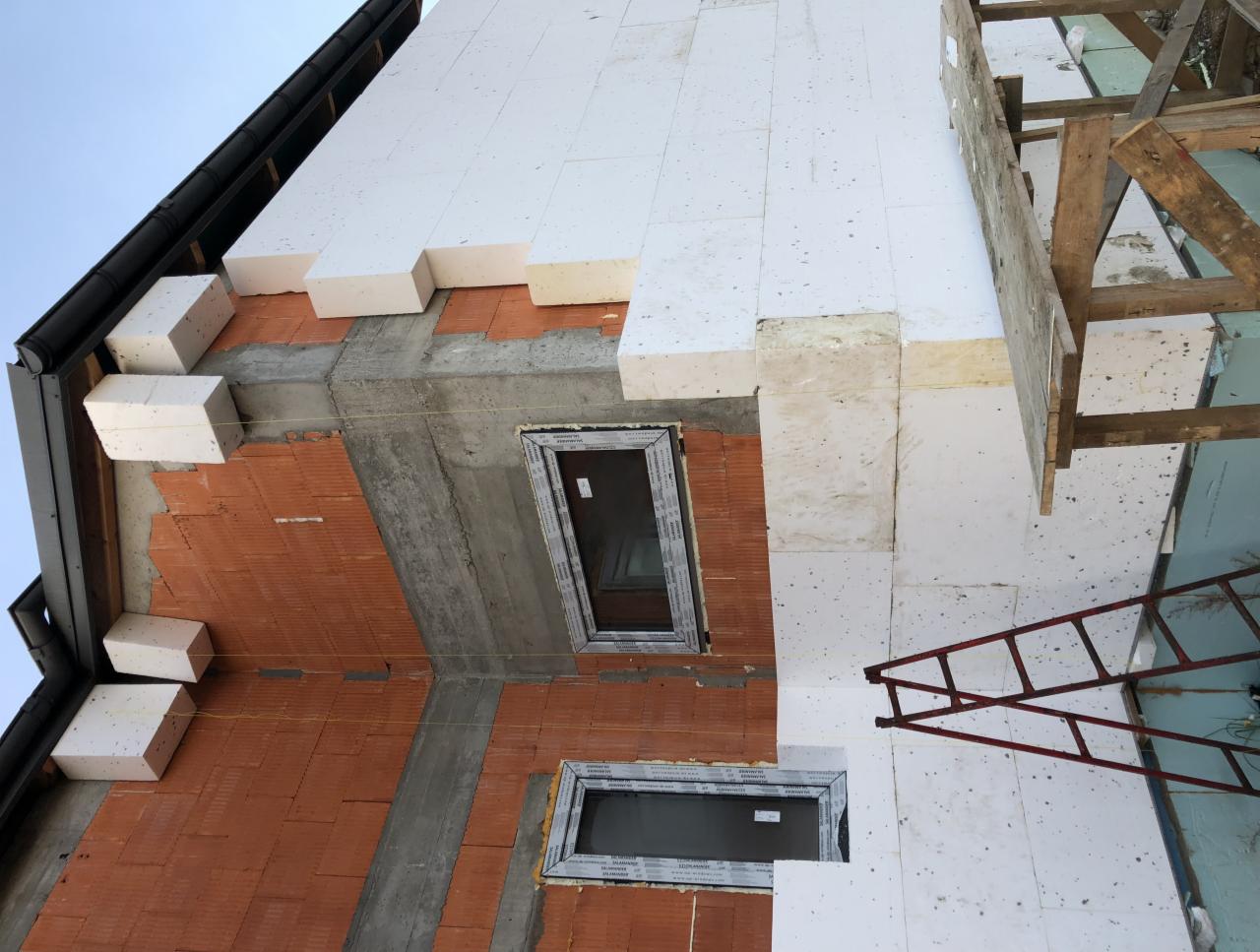 Fragment elewacji budynku w trakcie ocieplania styropianem, widoczne okna, rusztowanie i fragment dachu z rynną.