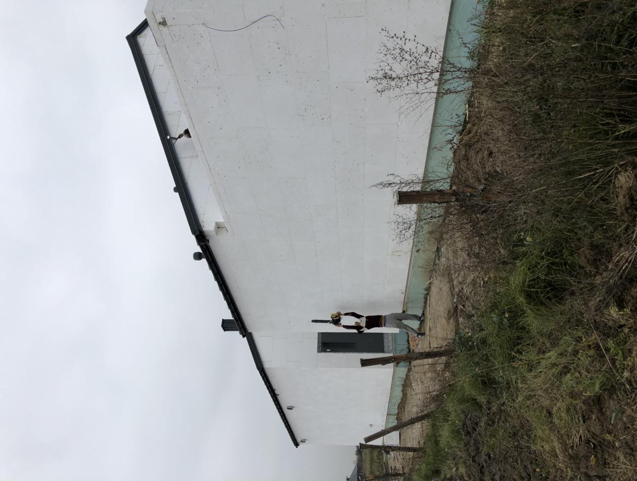 Pracownik w kombinezonie natryskuje białą farbę na ocieploną styropianem elewację budynku z widoczną czarną rynną i małym oknem, otoczoną zaniedbanym terenem z chwastami i drewnianymi słupkami.