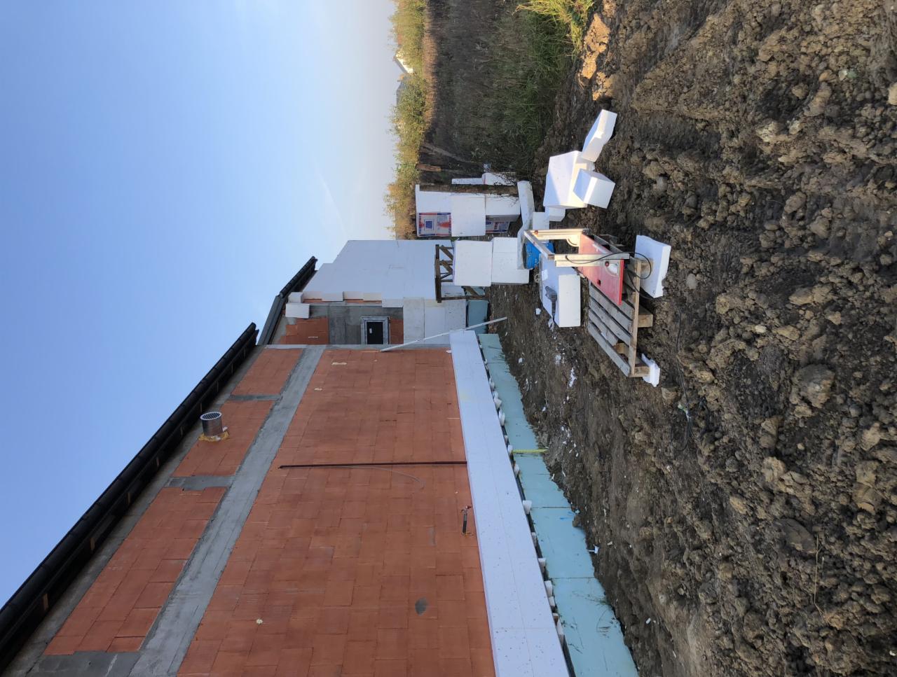 Budynek w trakcie ocieplania elewacji: widoczne płyty izolacyjne na ścianie, stos styropianu, narzędzie do cięcia styropianu i odkryty teren wokół budynku.