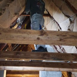 Arkbud - Pracownik w trakcie prac wykończeniowych na poddaszu, widoczne drewniane belki konstrukcyjne, siatka mocująca oraz ocieplenie, w tle widoczne pomieszczenie ze ścianami otynkowanymi na szaro i oknem...