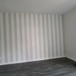 Pokój po remoncie: ściana w pionowe, szaro-białe pasy, szare panele podłogowe, biała szafka z przeszklonymi drzwiczkami oraz drabina malarska przy ścianie.