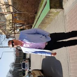 Arkbud - Mężczyzna w okularach przeciwsłonecznych, w jasnej marynarce i różowej koszuli, stoi na chodniku z czerwonej kostki brukowej, rzucając cień.