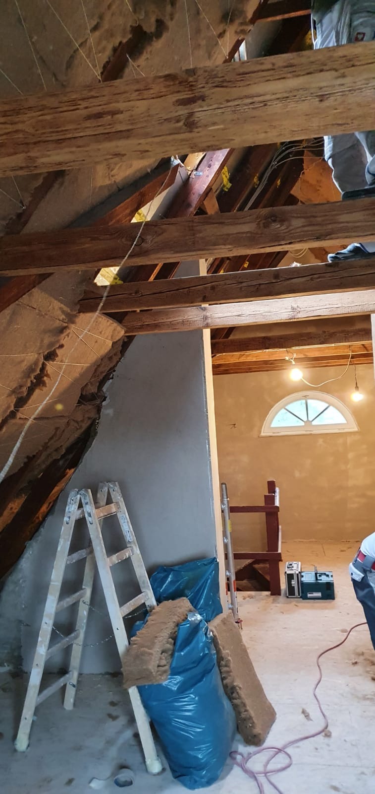Poddasze w trakcie prac wykończeniowych: ocieplona skośna ściana z widoczną konstrukcją drewnianą, drabina oparta o ścianę, worki z materiałem izolacyjnym, fragment pomieszczenia z oknem w tle.