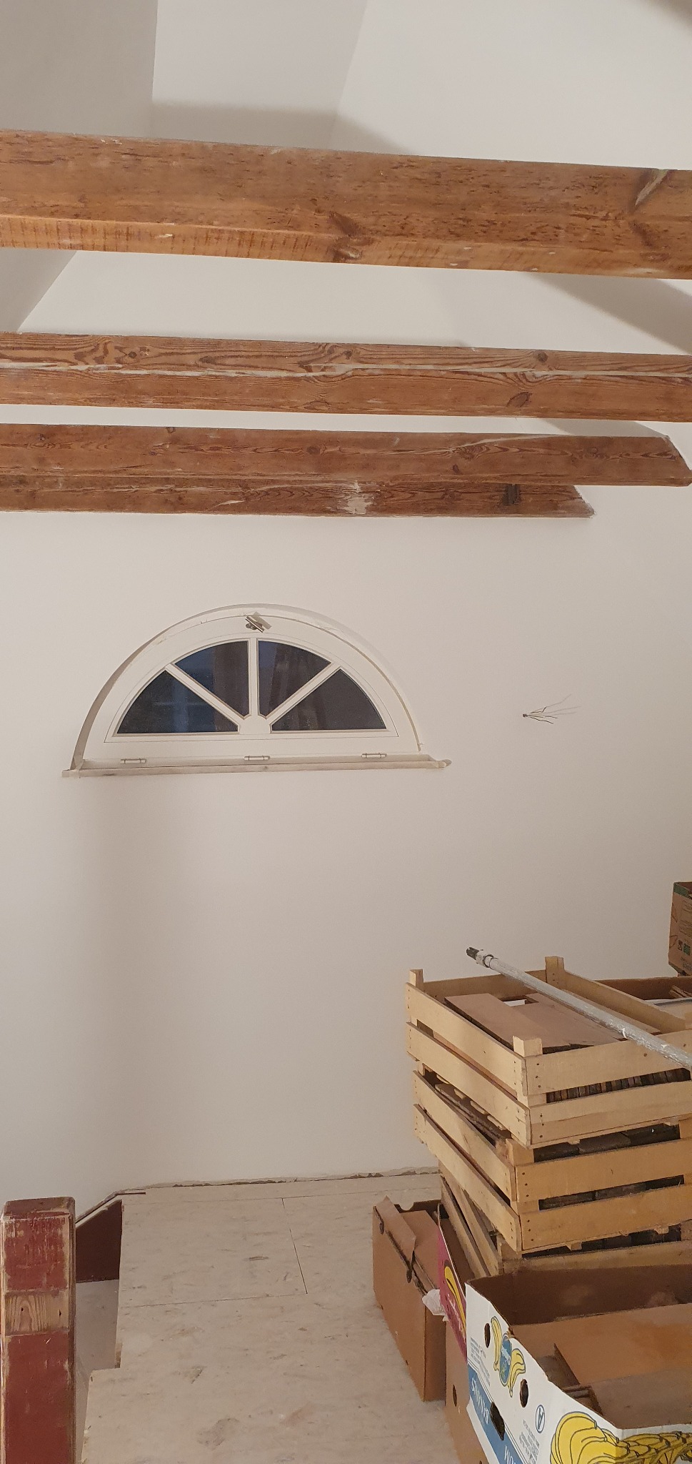 Wnętrze w trakcie prac wykończeniowych: widoczne drewniane belki stropowe, półokrągłe okno, przygotowane podłoże pod panele oraz stos skrzynek i kartonów.
