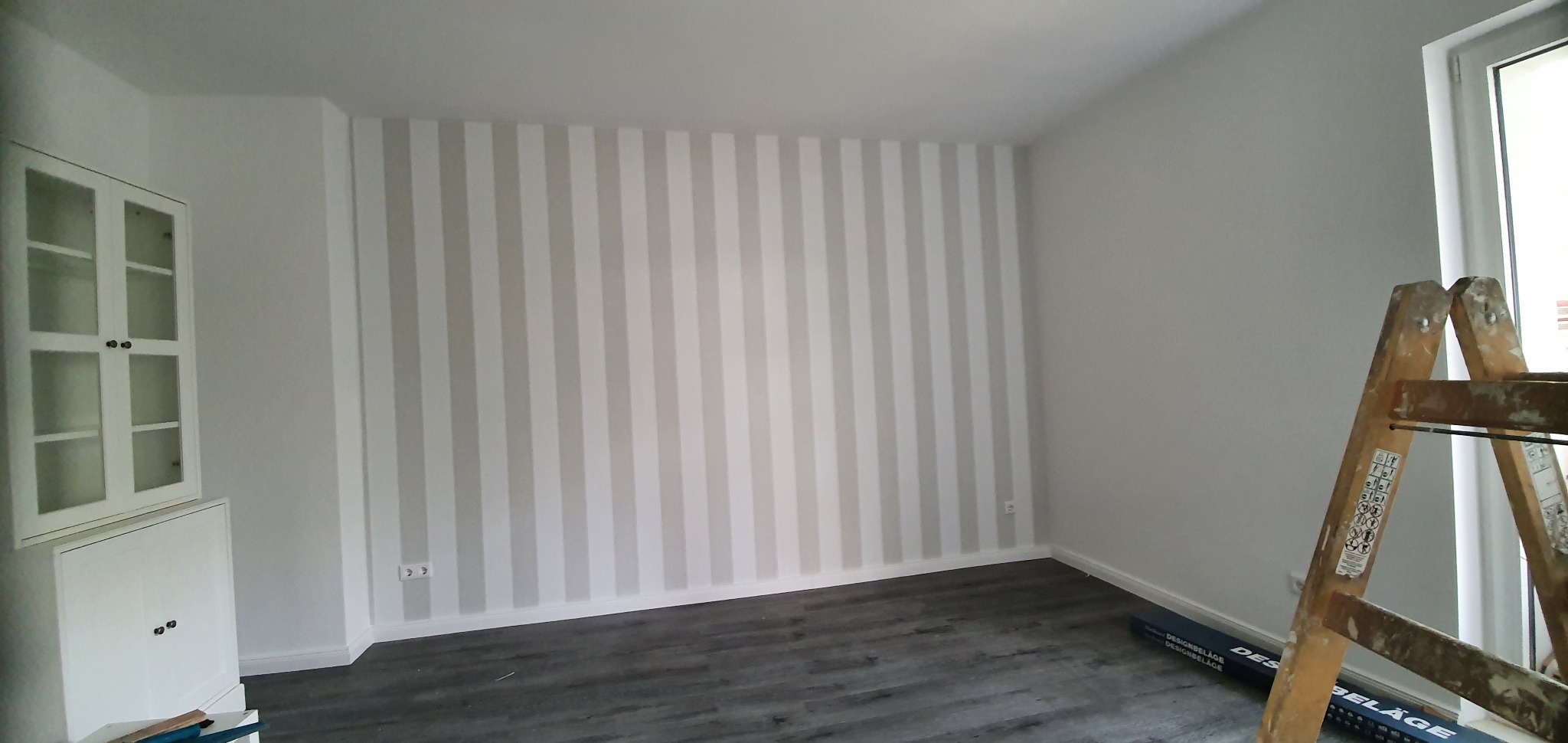 Pokój po remoncie: ściana w pionowe, szaro-białe pasy, szare panele podłogowe, biała szafka z przeszklonymi drzwiczkami oraz drabina malarska przy ścianie.