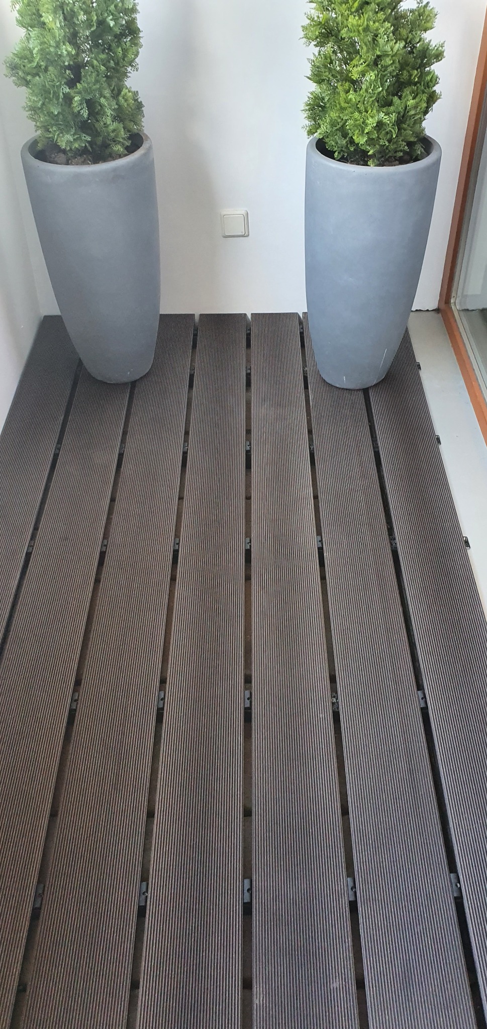 Balkon wyłożony brązowymi panelami podłogowymi z tworzywa sztucznego, pomiędzy którymi widoczne są plastikowe łączniki. Dwie szare, wysokie donice z zielonymi roślinami stoją przy ścianach po obu...