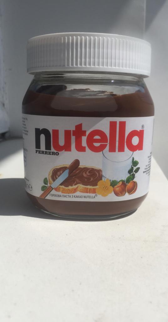 Słoik kremu czekoladowo-orzechowego Nutella, z widoczną etykietą, na której przedstawiono tost posmarowany kremem, szklankę mleka, orzechy laskowe i żółty kwiat.