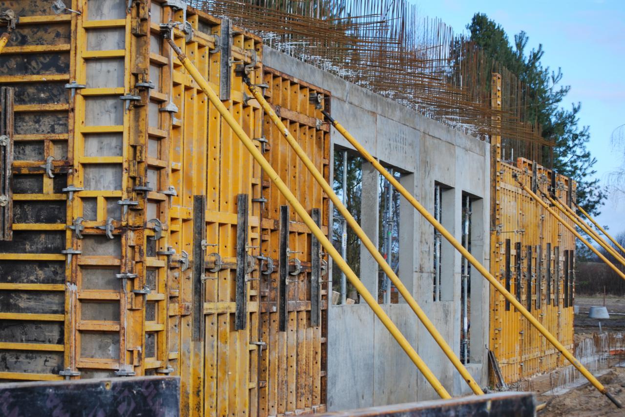 Szare betonowe fundamenty budynku z oknami w trakcie budowy, z żółtym szalunkiem i metalowym zbrojeniem.