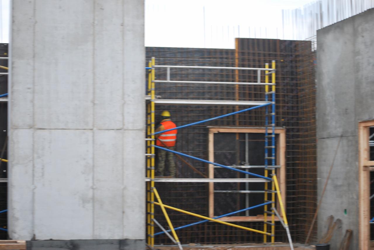 Pracownik w pomarańczowej kamizelce i żółtym kasku na rusztowaniu przy szalunku zbrojeniowym, widoczne betonowe elementy konstrukcyjne.