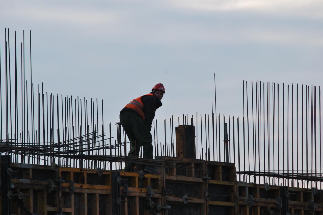 Pracownik w kasku i kamizelce odblaskowej pochylony nad szalunkiem z wystającymi prętami zbrojeniowymi, widok z dołu na tle pochmurnego nieba.