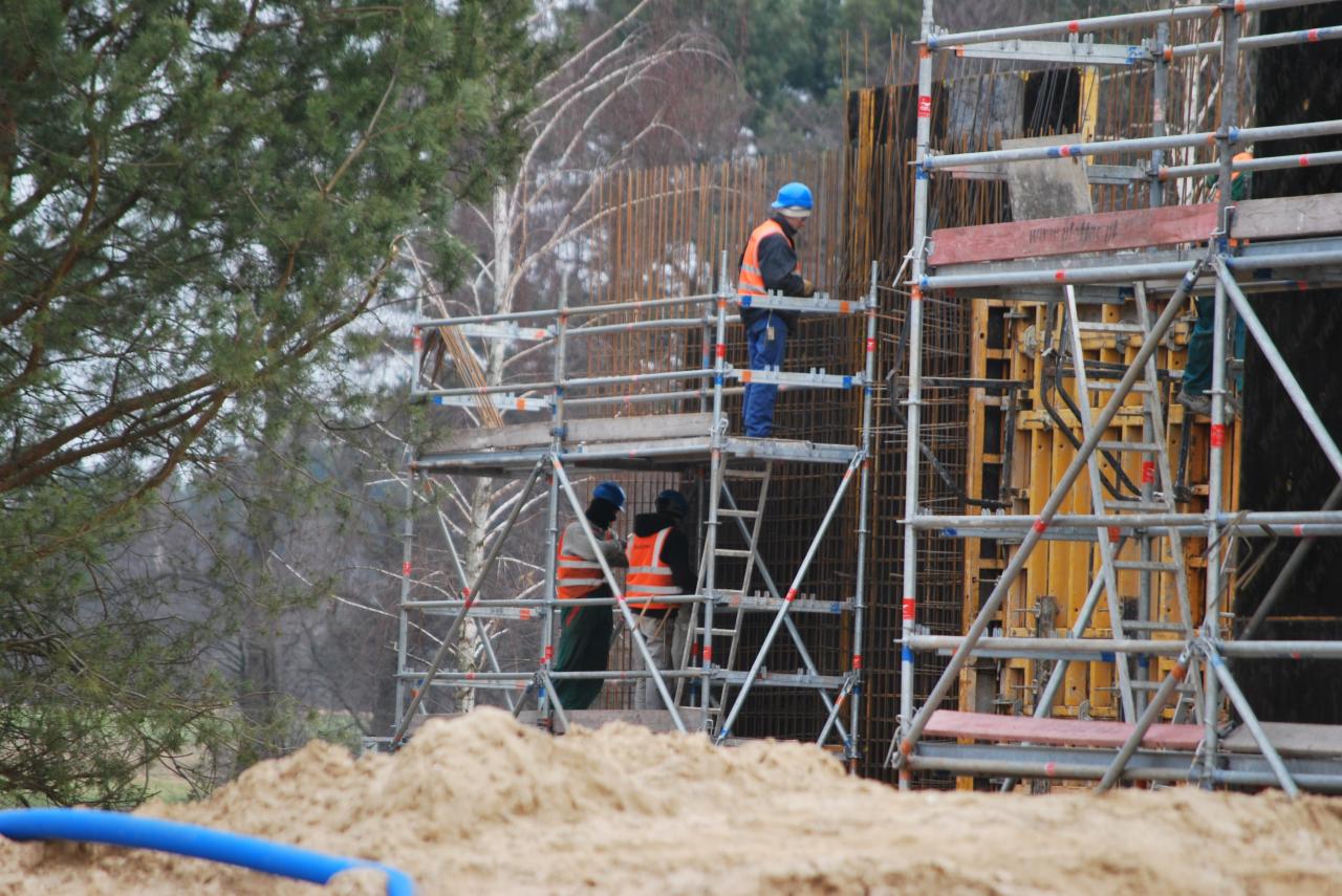 Dwóch pracowników w kaskach i kamizelkach odblaskowych na rusztowaniu przy szalunku zbrojeniowym, widoczny fragment niebieskiej rury i piasek w tle drzewa.