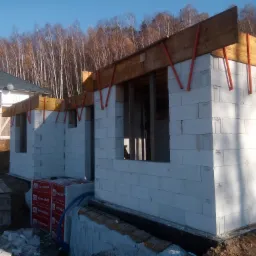 Wznoszenie ścian parteru domu z betonu komórkowego Termobet, widoczne drewniane szalunki okienne i zbrojenie pionowe, w tle drzewa i fragment gotowego domu.