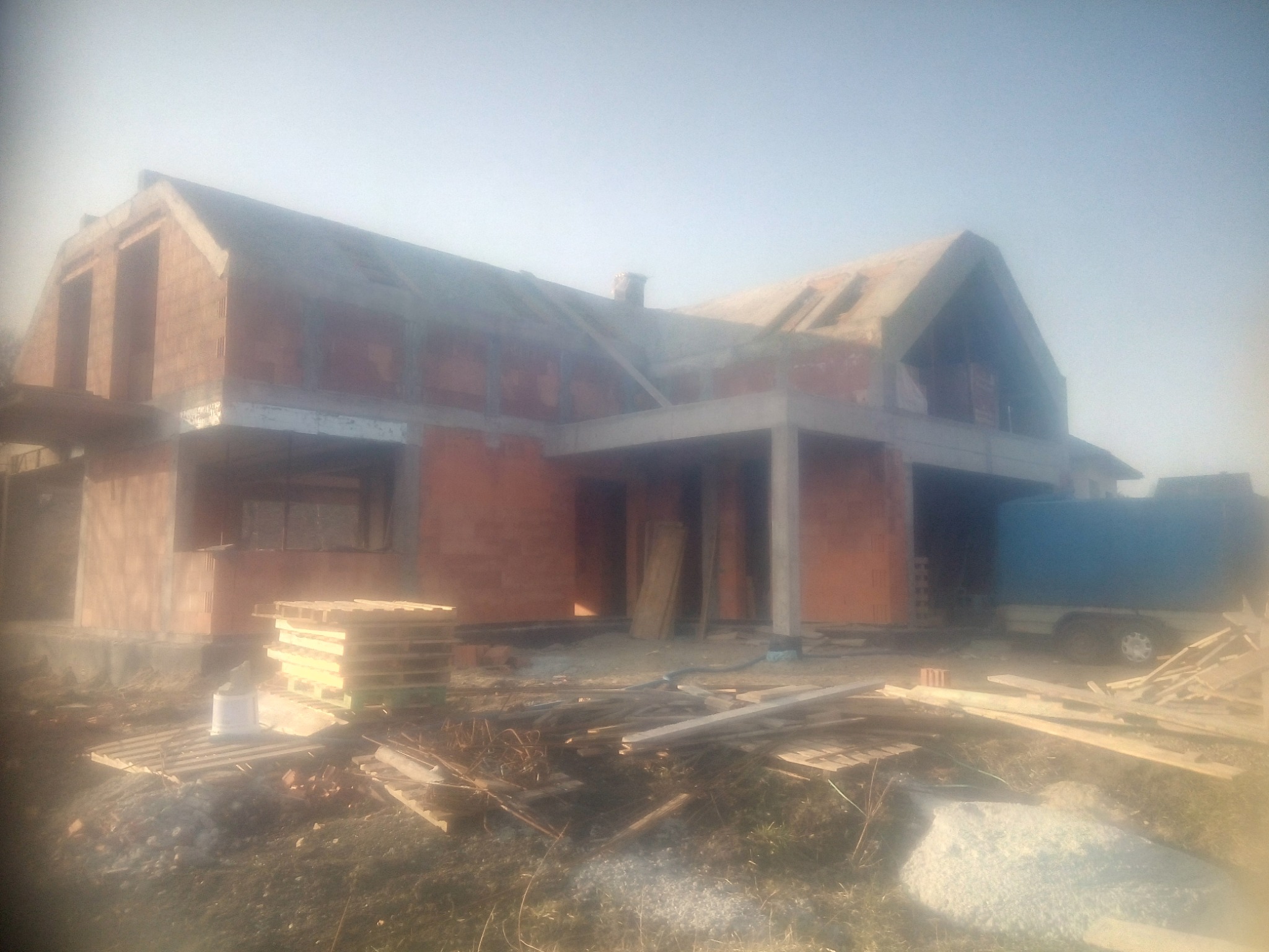 Niedokończony dom z czerwonej cegły w trakcie budowy, widoczne palety i materiały budowlane, zarys niebieskiej przyczepy w tle, pochmurne niebo.