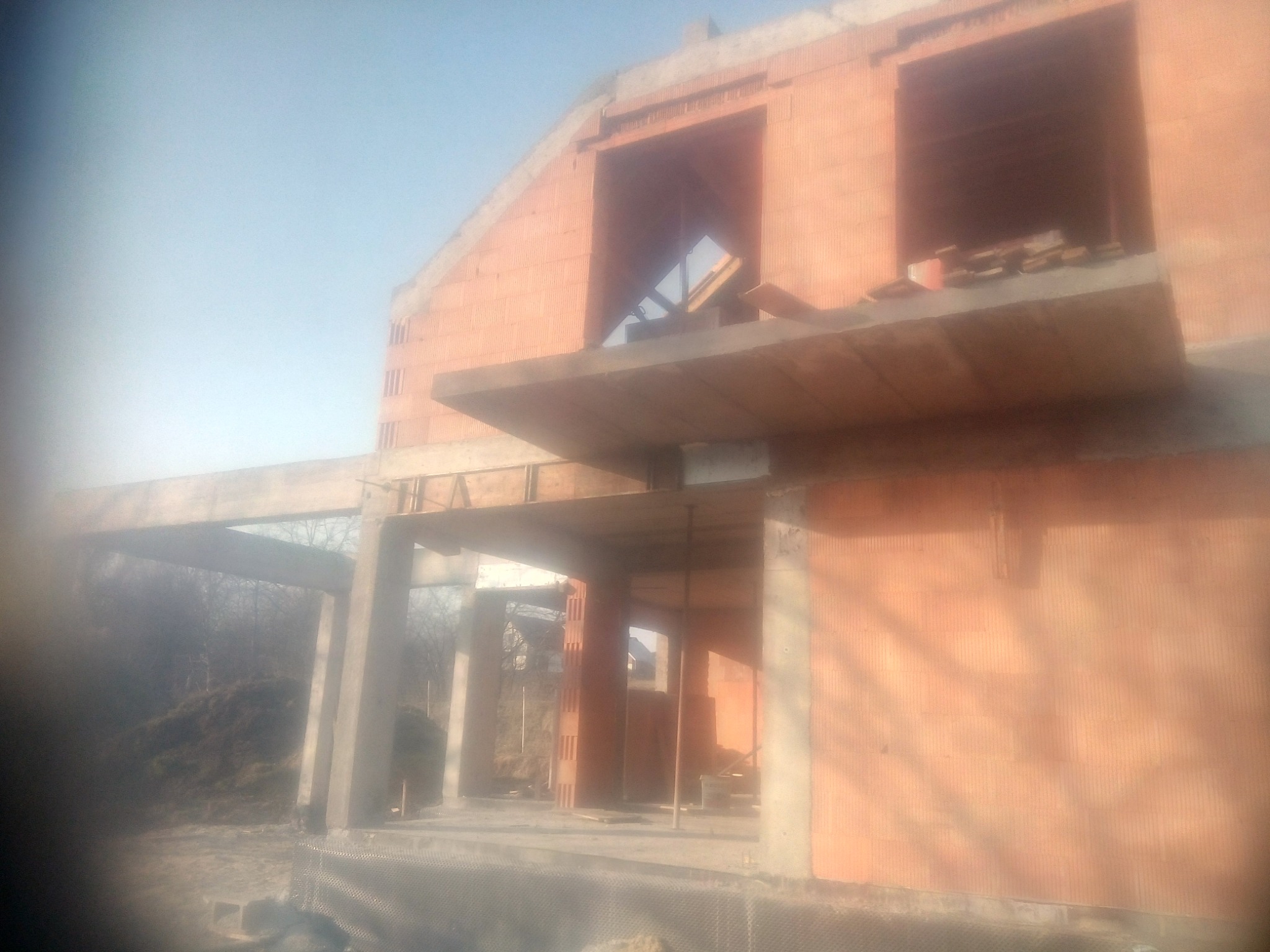 Surowy stan domu murowanego z betonu komórkowego, widoczne otwory okienne i betonowe nadproża, w tle drzewa. Budowa w trakcie realizacji.