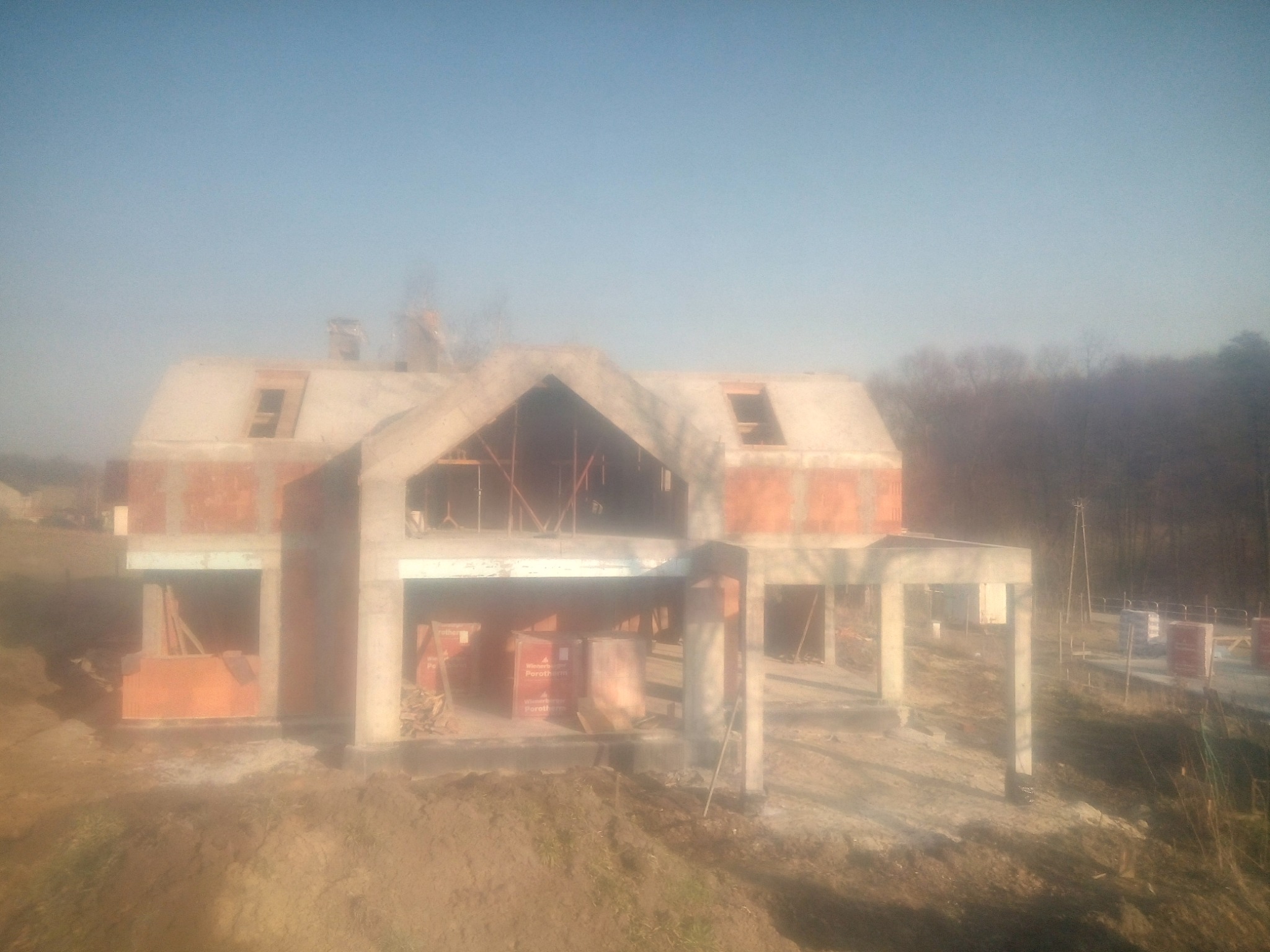 Surowy stan domu z betonu komórkowego w trakcie budowy, z widocznymi otworami okiennymi i zarysami dachu, na tle zalesionego terenu w mglisty dzień.