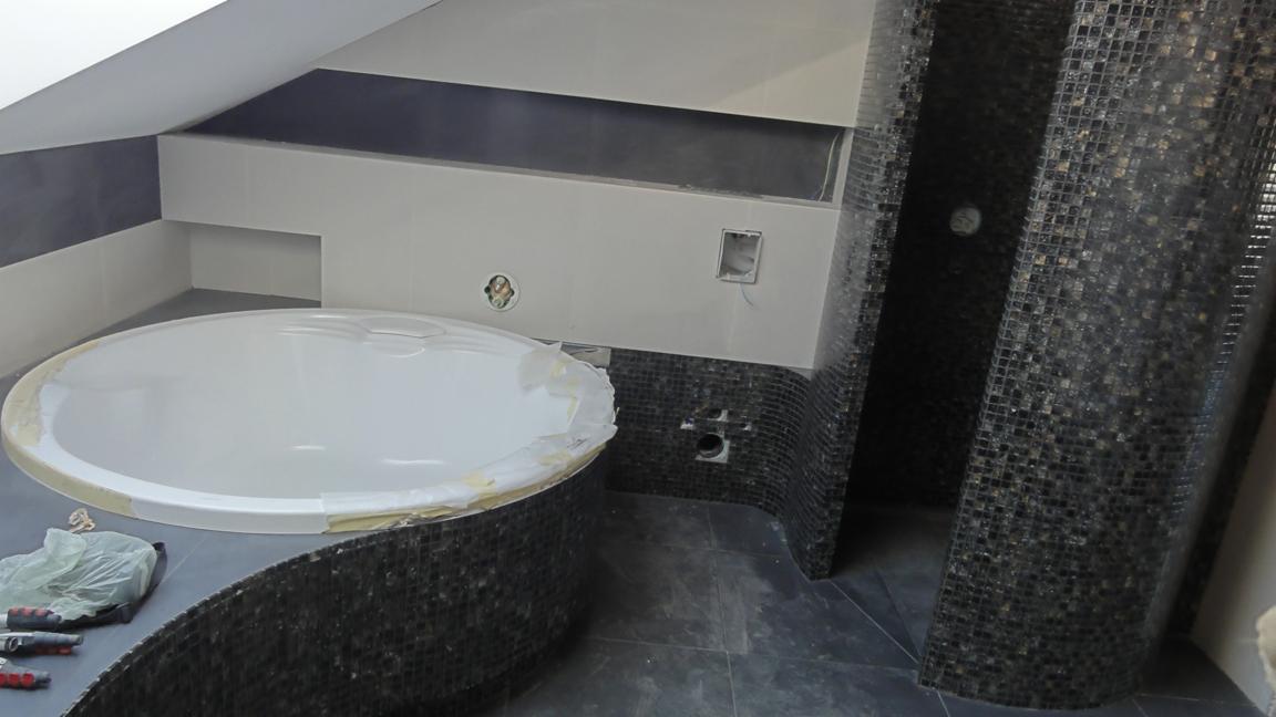Niedokończona łazienka w trakcie remontu: okrągła wanna z taśmą zabezpieczającą, ściany wyłożone mozaiką i płytkami, widoczne narzędzia i instalacje.